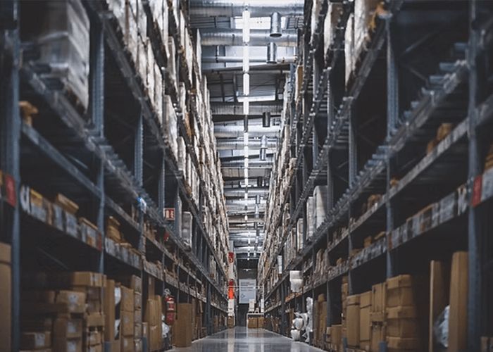 A long, narrow aisle in a large warehouse with tall shelves packed full of cardboard boxes.